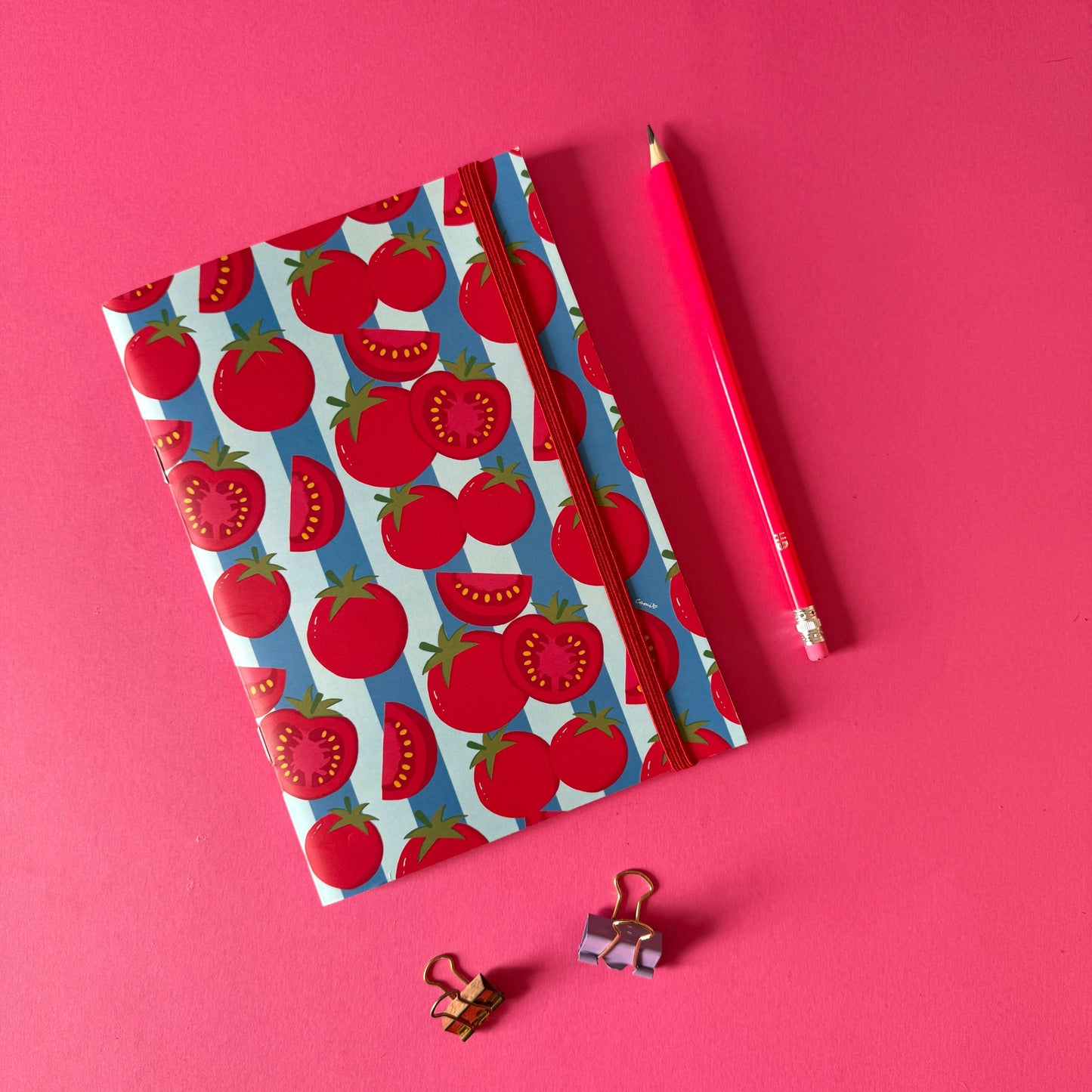 Libreta Frutalia Tomates con diseño de tomates rojos sobre fondo azul, lápiz y clips en color encima de una superficie rosa.