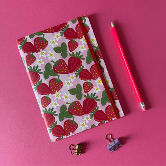 Libreta Frutalia Frutillas con diseño de fresas sobre fondo rosa, acompañada de lápiz y clips.