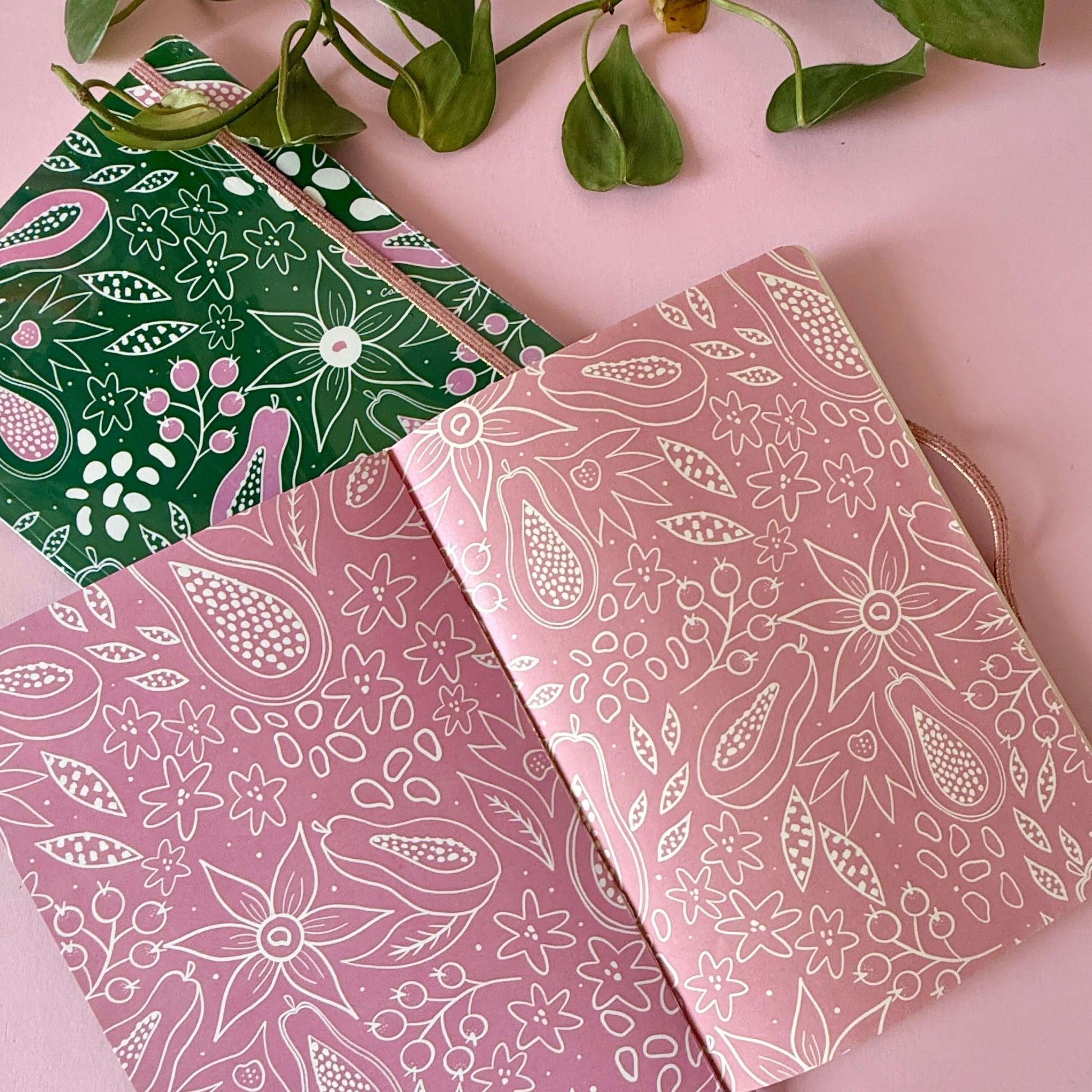 Libreta Flores verde y otras libretas con diseños florales sobre una superficie rosa.